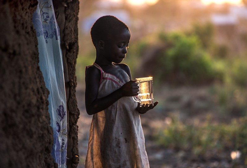 South Sudanese refugee Tabu Ruth brings water to her father, outside the family’s new home in Bidi Bidi camp. Photo : Tommy Trenchard